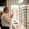 Man in a white shirt trying on glasses in a stylish eyewear store display with various styles.